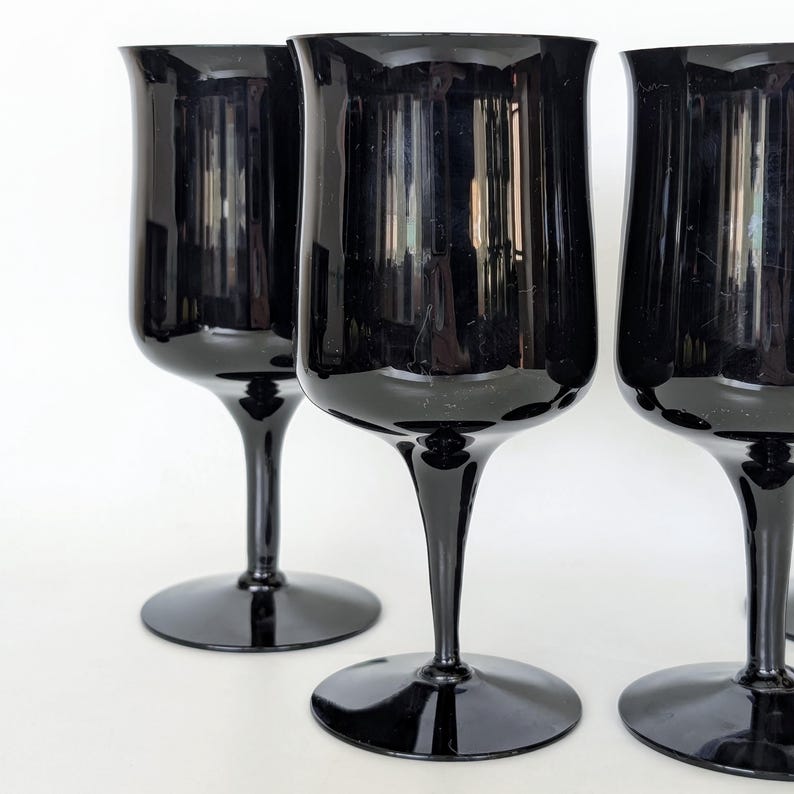 May include: Four black glass wine glasses with a wide bowl and a long, thin stem. The glasses are arranged in a row, with the front glass in focus.