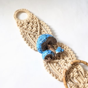 May include: A macrame plant hanger with a beige woven design, featuring a cluster of blue ceramic mushrooms. The mushrooms have a speckled glaze and brown stems. The hanger includes a wooden dowel and a loop for hanging.