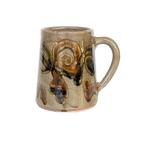 May include: A handmade ceramic mug with a tan base and a large handle. The mug features a decorative pattern of brown, green, and black swirls and shapes. The mug is suitable for hot or cold beverages.