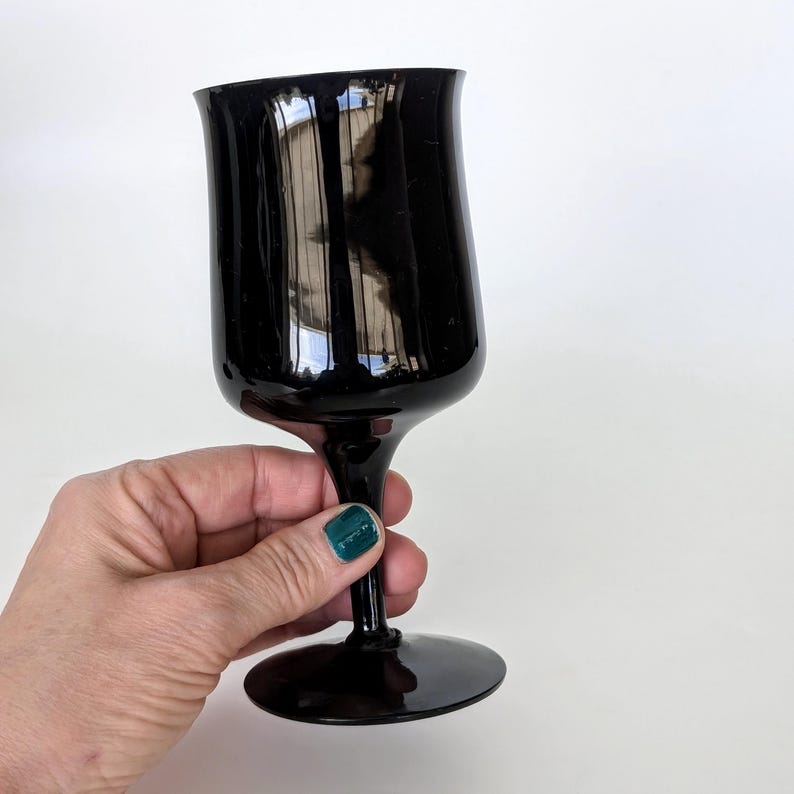 May include: A black glass wine goblet with a tall, slender stem and a wide, round bowl. The goblet is held in a hand with a green nail polish.