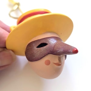May include: A decorative figurine of a character with a yellow hat, a burgundy mask, and a long nose with a red tip. The figurine has a peach-colored face with a small smile and a rosy cheek. The hat has a red band.