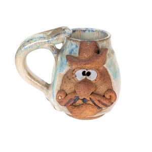 May include: A ceramic mug with a cowboy face design. The mug features a tan and brown color scheme with blue accents. The handle is integrated into the design. The face has a hat, eyes, nose, and mustache.