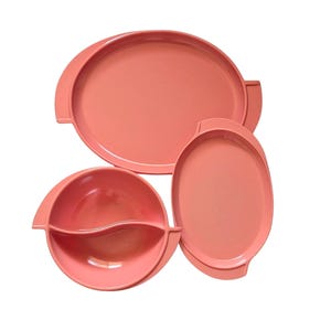May include: A set of three coral-colored serving dishes including one large oval platter, one smaller oval platter, and one round bowl divided into two sections by a central curved wall. Each piece features integrated side handles for easy gripping.