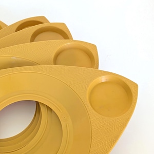 May include: A stack of mustard yellow plastic plates, each featuring a textured surface, a large central circular cutout, and a smaller circular indentation on one side. The plates are arranged in a fanned-out, overlapping formation.