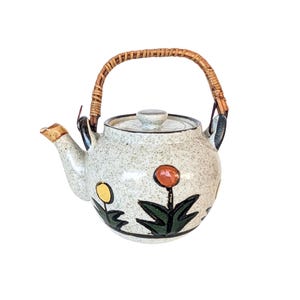 May include: A white ceramic teapot with a speckled glaze and a brown wicker handle. The teapot is decorated with green leaves and yellow and orange flowers.