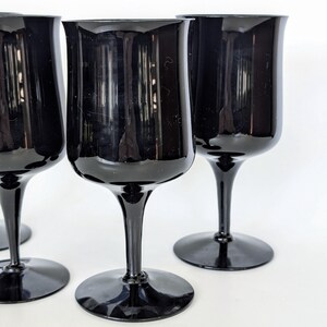 May include: Four black glass wine goblets with a wide bowl and a long stem. The goblets are arranged in a row on a white surface.