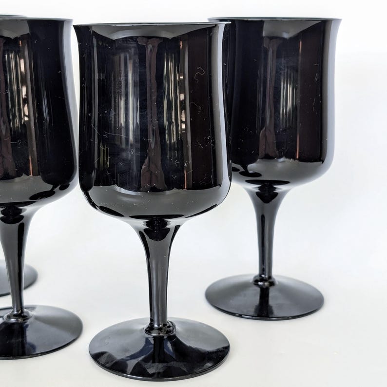 May include: Four black glass wine goblets with a wide bowl and a long stem. The goblets are arranged in a row on a white surface.