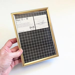 May include: A gold-colored rectangular picture frame, labeled "Ripa 13 x 18 (5 x 7 in)" with a black and white grid pattern visible through the glass. The frame is held by a hand against a white background.