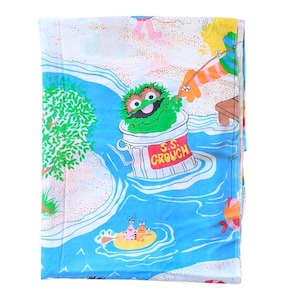 May include: A vintage Sesame Street flat sheet featuring a colorful beach scene. The design includes Oscar the Grouch in a trash can labeled "S.S. Crouch", a blue river, and other Sesame Street characters. The sheet is primarily white and blue.