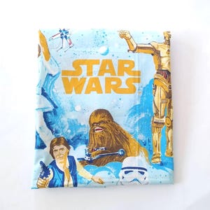May include: A light blue Star Wars themed bed sheet featuring the words "STAR WARS" in gold. The sheet has illustrations of Chewbacca, C-3PO, a Stormtrooper, and other characters from the movie.