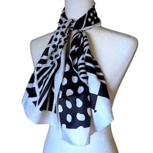 May include: A black and white patterned scarf draped over a mannequin. The scarf features various geometric designs, including polka dots, stripes, and angular shapes. The scarf is made of a lightweight material and is knotted at the neck.