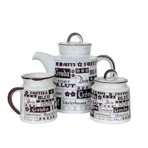 May include: A white ceramic tea set with brown and black lettering. The teapot, creamer, and sugar bowl are decorated with a pattern of cheese names, including Gouda, Brie, and Fontina.