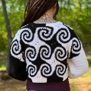 Peut inclure: Un pull en crochet noir et blanc avec un motif en spirale. Le pull est court et a des manches longues.