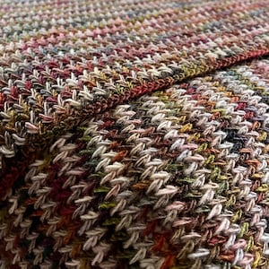 May include: A multi-colored knitted throw blanket with a chevron pattern in shades of brown, beige, pink, and green. The blanket is folded on a white bed.