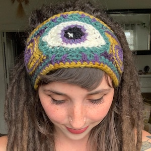May include: A crochet headband featuring a prominent central eye motif with a black pupil, white iris, and purple outline. The band is crafted with teal, mustard yellow, and purple yarn, showcasing intricate patterns that wrap around the head.
