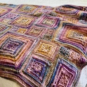 Peut inclure: Une couverture au crochet faite à la main, multicolore, avec un motif géométrique en losanges. La couverture présente des nuances de violet, d'or, de marron et de bleu. La couverture est drapée sur un lit blanc, créant une esthétique chaleureuse et accueillante.