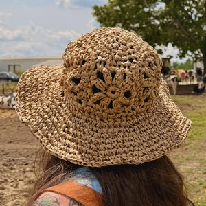 Puede incluir: Un sombrero de sol de paja tejida con ala ancha y un diseño floral en la corona. El sombrero es de color marrón claro y parece hecho a mano. La persona que lleva el sombrero tiene el pelo largo y castaño y un tatuaje en el brazo.