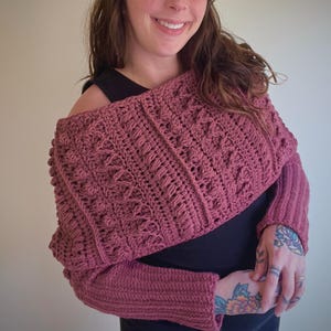 May include: A dusty rose crochet shrug with intricate stitch patterns. The off-the-shoulder design features long sleeves with ribbed cuffs. The handmade bolero is a cozy and stylish accessory, perfect for layering.