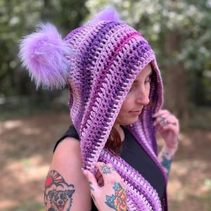 May include: A purple and pink striped crocheted hooded scarf with two fluffy pom-poms on the hood.