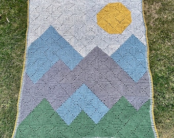 Könnte beinhalten: Eine gehäkelte Decke mit einem Berg- und Sonnenmotiv. Die Decke ist aus verschiedenen Blautönen, Grüntönen und Gelbtönen gefertigt. Die Decke hat einen gelben Rand.