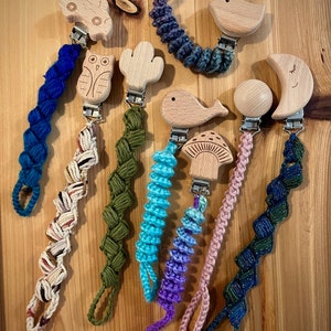 May include: A set of eight wooden baby teethers with crocheted straps in various colors. The teethers feature different shapes, including a car, owl, cactus, whale, mushroom, bird, moon, and a round shape.