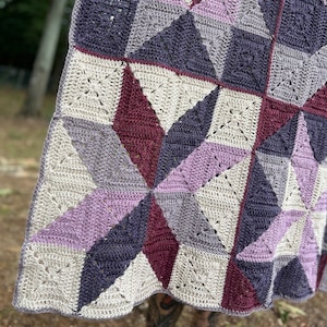 Peut inclure: Une couverture au crochet faite à la main avec un motif géométrique. La couverture présente des carrés et des triangles dans des tons de violet, lavande, gris et crème. La couverture est exposée à l'extérieur, avec un arrière-plan flou d'arbres.