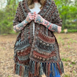 Pode incluir: Um xale de crochê marrom, verde e ferrugem com franjas. O xale tem dois bolsos grandes e um design texturizado.