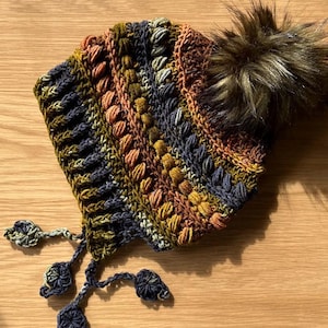 Op de afbeelding: Een handgemaakte gehaakte muts in een mix van kleuren, waaronder olijfgroen, marineblauw en perzik. De muts heeft een textuurpatroon en een grote, pluizige bruine pompon bovenop. Decoratieve kwastjes hangen aan de zijkanten.