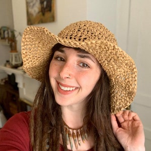 May include: A woman with long brown dreadlocks is wearing a wide-brimmed straw hat, a maroon long-sleeved shirt, and a necklace with brown and blue stones. She has a nose ring and a tattoo on her arm.