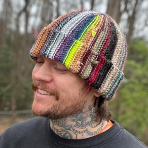 Puede incluir: Una persona lleva una boina de rayas de colores con un esquema de colores marrón, gris, morado, rojo, verde y amarillo. La boina está hecha a ganchillo y tiene un ajuste holgado.