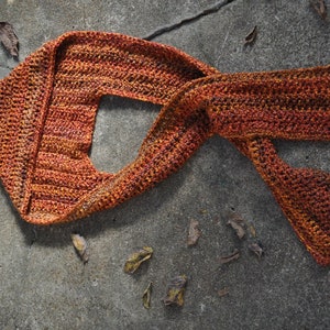 May include: A crocheted scarf in shades of orange and brown, lying on a gray concrete surface.