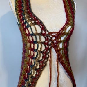 May include: A sleeveless crochet vest featuring an open, airy stitch pattern in a variegated color palette of deep red, olive green, dark blue, and brown. The garment has a V-neckline and includes long, thin yarn ties at the center front for closure.