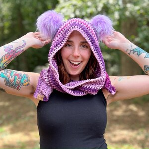 May include: A purple and pink hooded scarf with fuzzy earmuffs. The scarf is crocheted and has a gradient of color from purple to pink. The earmuffs are made of fluffy purple fur.