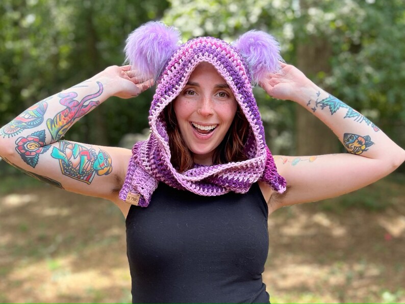 May include: A purple and pink hooded scarf with fuzzy earmuffs. The scarf is crocheted and has a gradient of color from purple to pink. The earmuffs are made of fluffy purple fur.