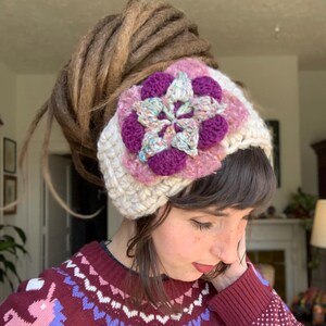 May include: A thick, cream-colored crocheted headband featuring a large, multi-layered floral applique. The flower is composed of deep purple, light pink, and variegated pastel yarn petals, creating a textured, layered appearance on the band.