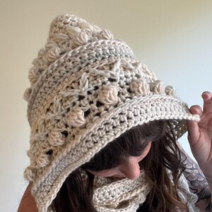 Op de afbeelding: Een cr&egrave;mekleurige gehaakte capuchon- en sjaalset. De capuchon heeft een puntige bovenkant en is versierd met getextureerde, verhoogde bobbles. De sjaal heeft een bijpassende cr&egrave;mekleur en heeft een vergelijkbaar open weefpatroon. De set lijkt handgemaakt.