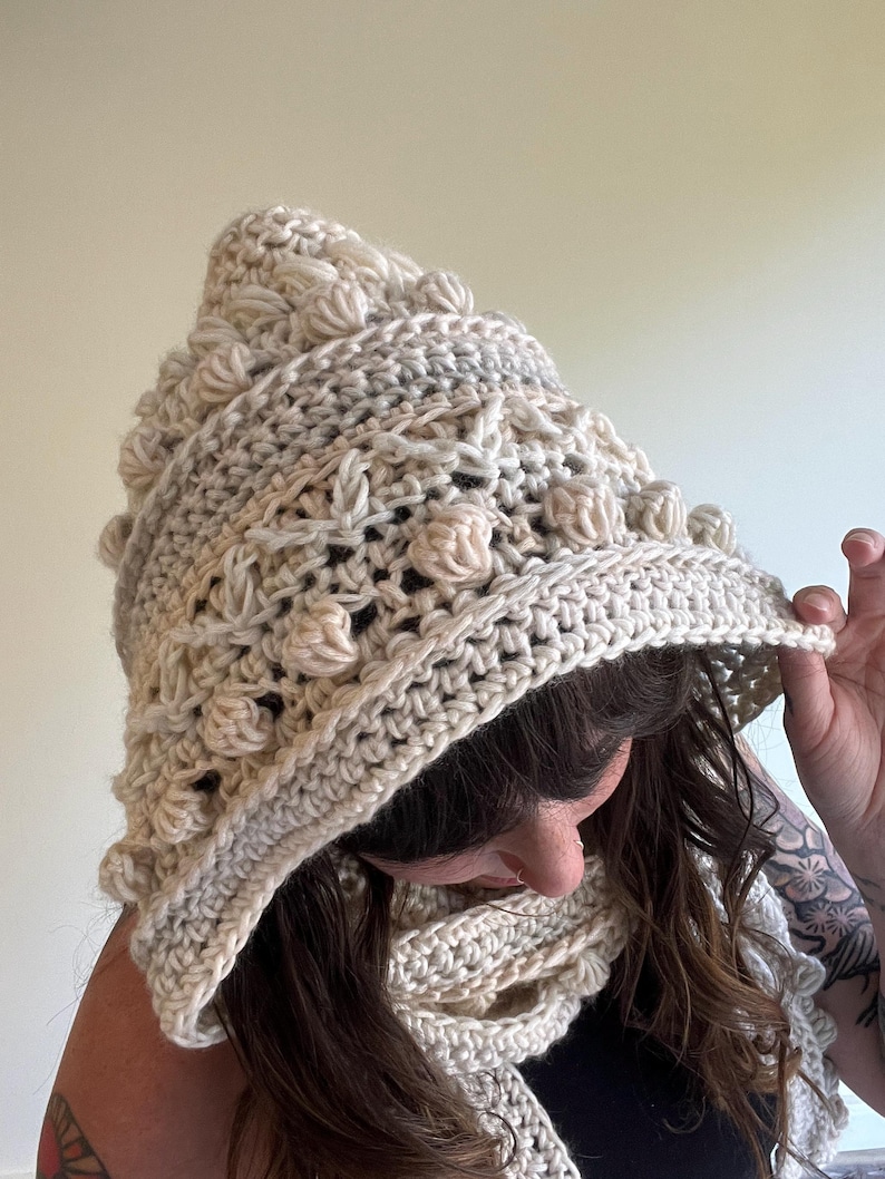 Op de afbeelding: Een cr&egrave;mekleurige gehaakte capuchon- en sjaalset. De capuchon heeft een puntige bovenkant en is versierd met getextureerde, verhoogde bobbles. De sjaal heeft een bijpassende cr&egrave;mekleur en heeft een vergelijkbaar open weefpatroon. De set lijkt handgemaakt.
