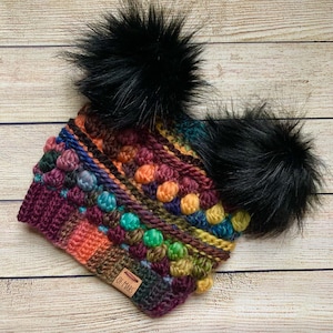 Puede incluir: Un gorro de punto de crochet colorido con un hilo multicolor y un punto de bobble texturizado. El gorro tiene dos pompones de piel sintética negra en la parte superior y una etiqueta de cuero marrón que dice "Of Mars".