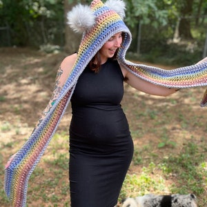May include: A rainbow striped crochet hooded scarf with white pom poms on the hood. The scarf is being worn by a person with tattoos on their arms.