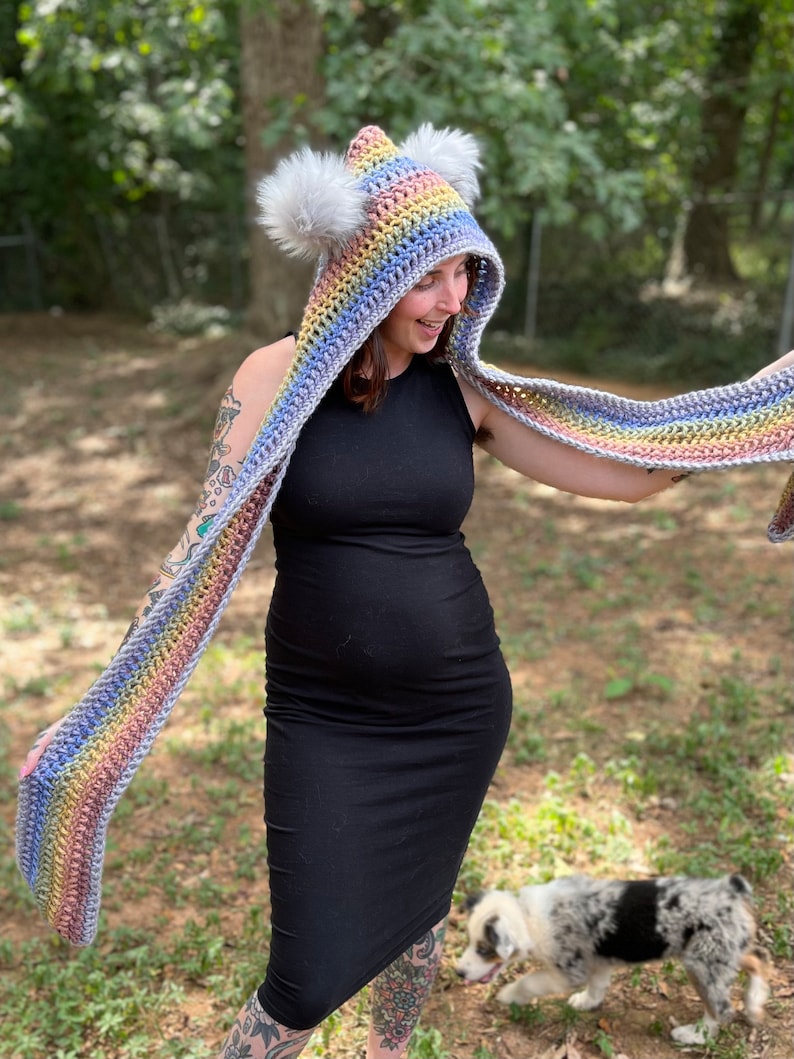 May include: A rainbow striped crochet hooded scarf with white pom poms on the hood. The scarf is being worn by a person with tattoos on their arms.