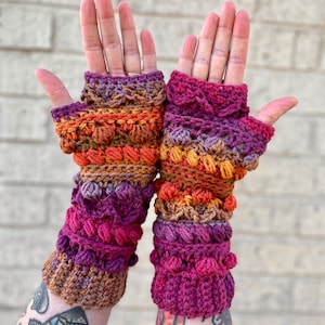 May include: A pair of crocheted fingerless gloves in a multi-colored ombre pattern of purple, pink, orange, and brown. The gloves have a textured design with small bobbles.