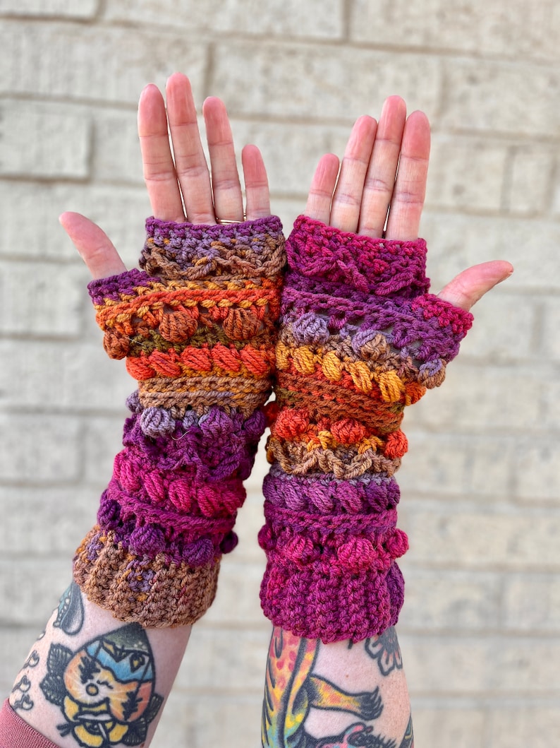 May include: A pair of crocheted fingerless gloves in a multi-colored ombre pattern of purple, pink, orange, and brown. The gloves have a textured design with small bobbles.