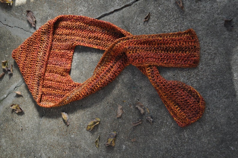 May include: A crocheted scarf in shades of orange and brown, lying on a gray concrete surface.