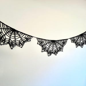 Op de afbeelding: Zwarte spinnenweb slinger, perfect voor Halloween-decoraties. De slinger heeft drie spinnenweb ontwerpen die zijn verbonden door een dun zwart touwtje. De spinnenwebben hebben een gedetailleerd, ingewikkeld ontwerp.