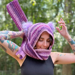 May include: A purple and pink striped crocheted cowl with fuzzy ear-like attachments. The cowl is being worn by a person with tattoos on their arms.
