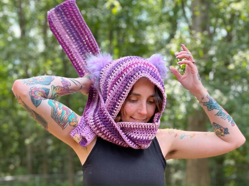 May include: A purple and pink striped crocheted cowl with fuzzy ear-like attachments. The cowl is being worn by a person with tattoos on their arms.