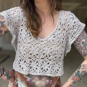 May include: A light gray, handmade crochet top with a V-neck and short, ruffled sleeves. The top features a floral pattern throughout the bodice and sleeves, showcasing the intricate openwork design. This garment is a stylish addition to any wardrobe.