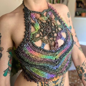 May include: A multi-colored crochet halter top with a large, openwork, crocheted design in the center. The top is made with a variety of shades of green, blue, purple, and brown yarn.