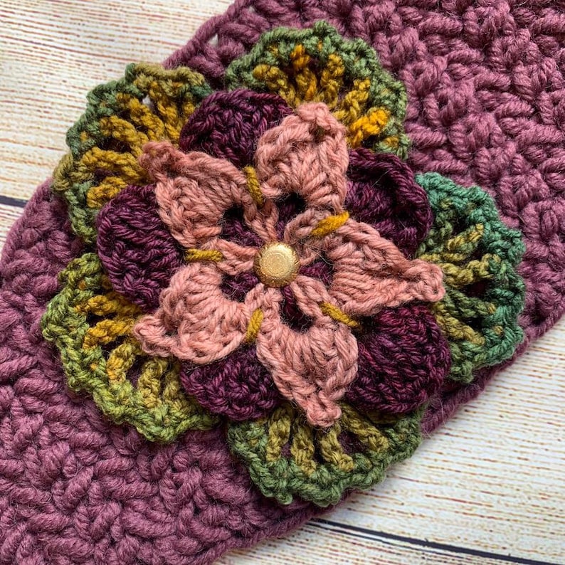 May include: A crochet headband in a deep plum color featuring a layered floral embellishment. The flower consists of petals in shades of dusty pink, dark purple, and olive green, centered with a small, round, metallic gold-toned button.