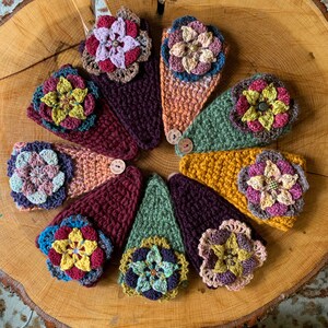 May include: A collection of eight triangular crochet headbands arranged in a circle. Each headband features a unique color combination of yarn and a large, multi-layered crocheted flower embellishment attached to the center with a small button.